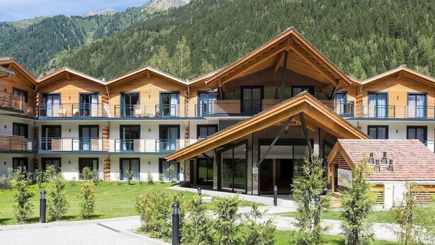 Een modern hotel met meerdere houten balkons, een centrale ingang en een bergachtige achtergrond.