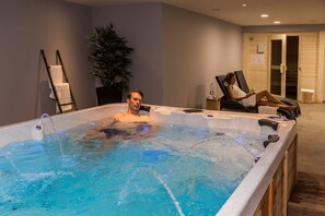 A man in a hot tub with a towel on the side, a plant, and a chair with a person reading in the background.