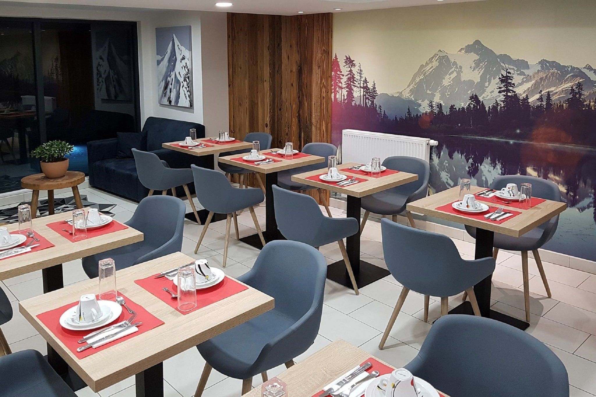 Un espace repas avec des tables dressées pour quatre personnes, des chaises bleues et une fresque représentant un paysage de montagne sur le mur.