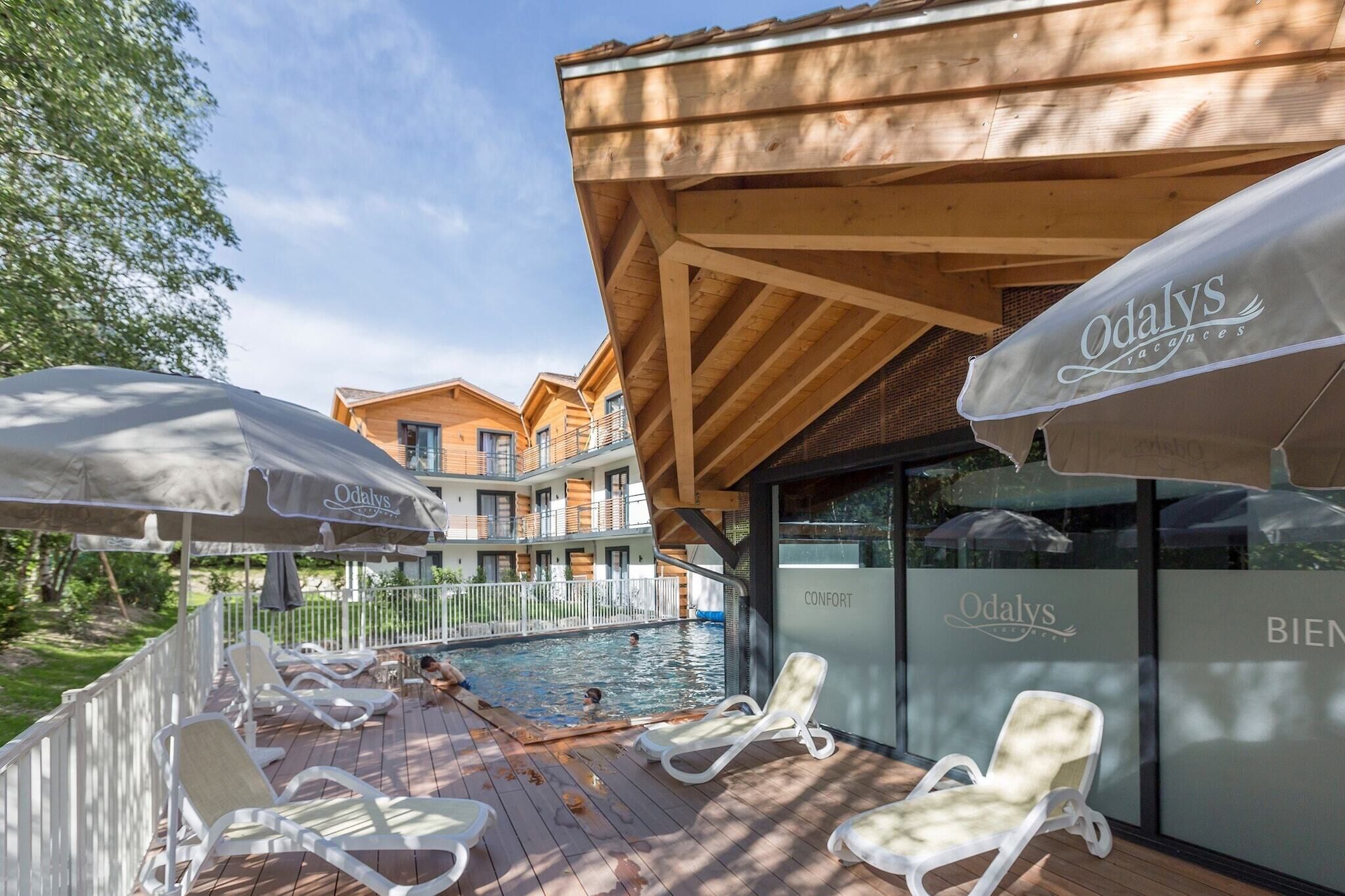 A poolside area with lounge chairs and umbrellas, a wooden deck, and a building with 'Odalys' branding.
