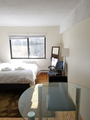 A hotel room with a bed, a chair, a glass table, and a window with a view of buildings. - Packard's Corner Apartments by Atlas (Boston)