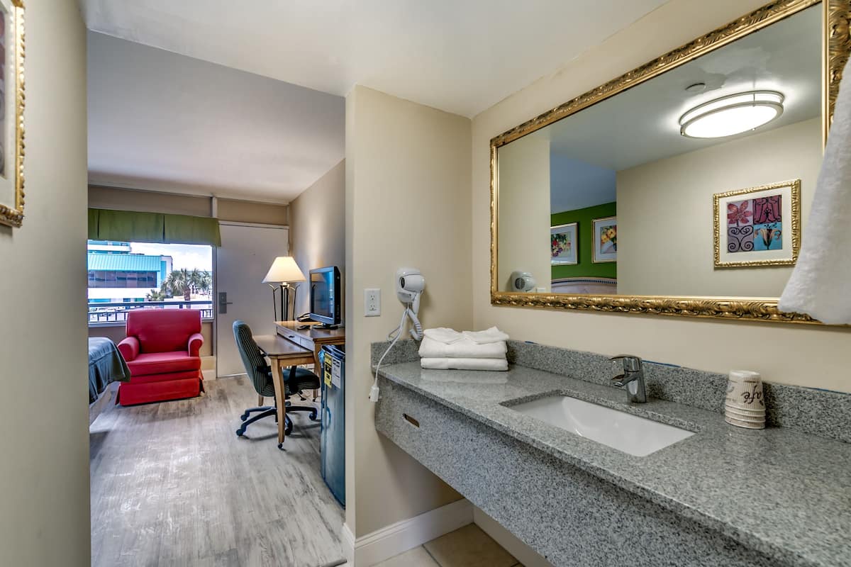 Une chambre d’hôtel avec un grand miroir, un lavabo sur un plan de travail en granit, une chaise rouge, un bureau avec une lampe et une vue sur un bâtiment à l’extérieur.