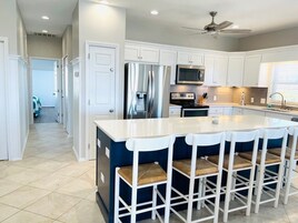 Una cocina moderna con armarios blancos, electrodomésticos de acero inoxidable, una isla central con taburetes altos y un ventilador de techo.