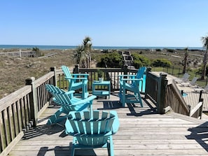 A wooden deck with turquoise Adirondack chairs and a small table, overlooking a beach and ocean.