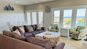 Ein Wohnzimmer mit Ecksofa, Couchtisch, zwei Sesseln, einem Esstisch mit Stühlen und großen Fenstern mit Blick auf den Strand.