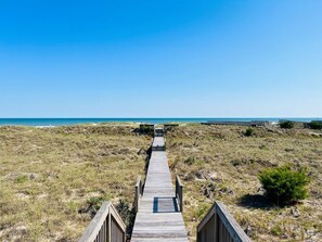 Un paseo marítimo de madera que conduce a una playa con dunas y un cielo azul despejado.