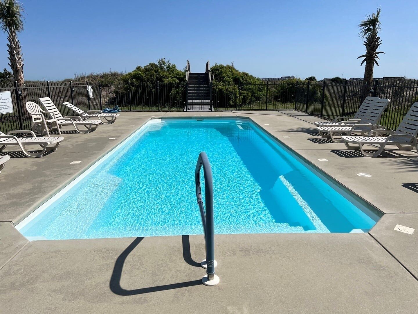 Une piscine rectangulaire dotée d’une rambarde métallique, entourée de chaises longues blanches et de palmiers.