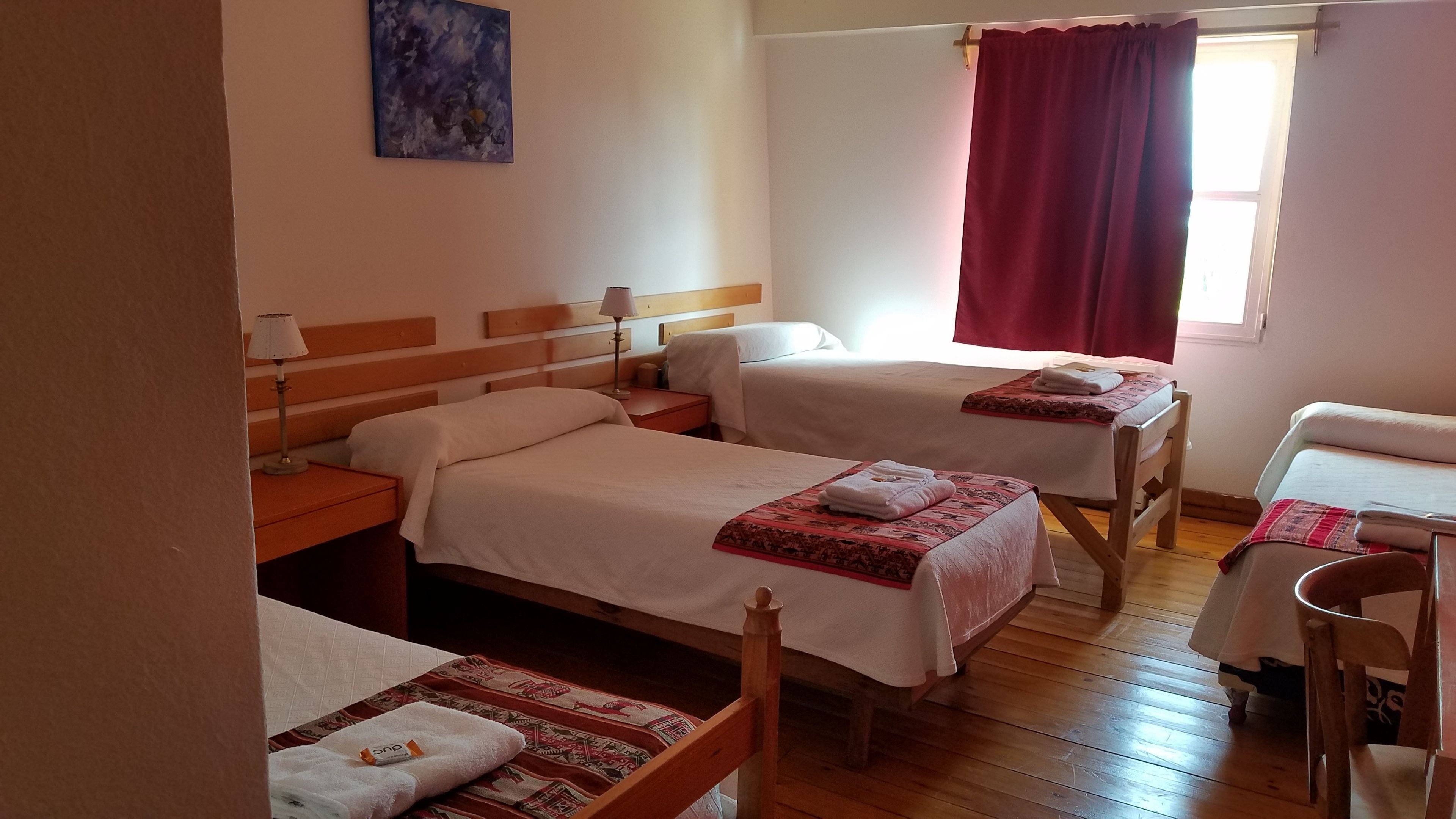 Une chambre d’hôtel avec deux lits simples, une tête de lit en bois, une table de chevet avec une lampe, une fenêtre avec des rideaux rouges et une petite table avec une chaise.