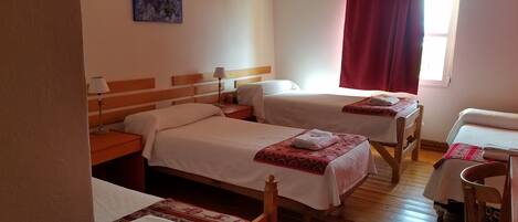 Une chambre d’hôtel avec deux lits simples, une tête de lit en bois, une table de chevet avec une lampe, une fenêtre avec des rideaux rouges et une petite table avec une chaise.