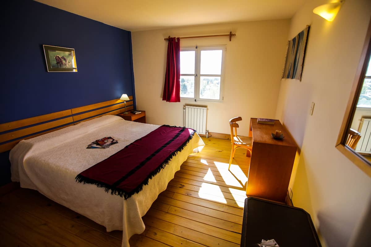 Un dormitorio con cama, suelo de madera, una ventana con cortinas, escritorio y radiador.