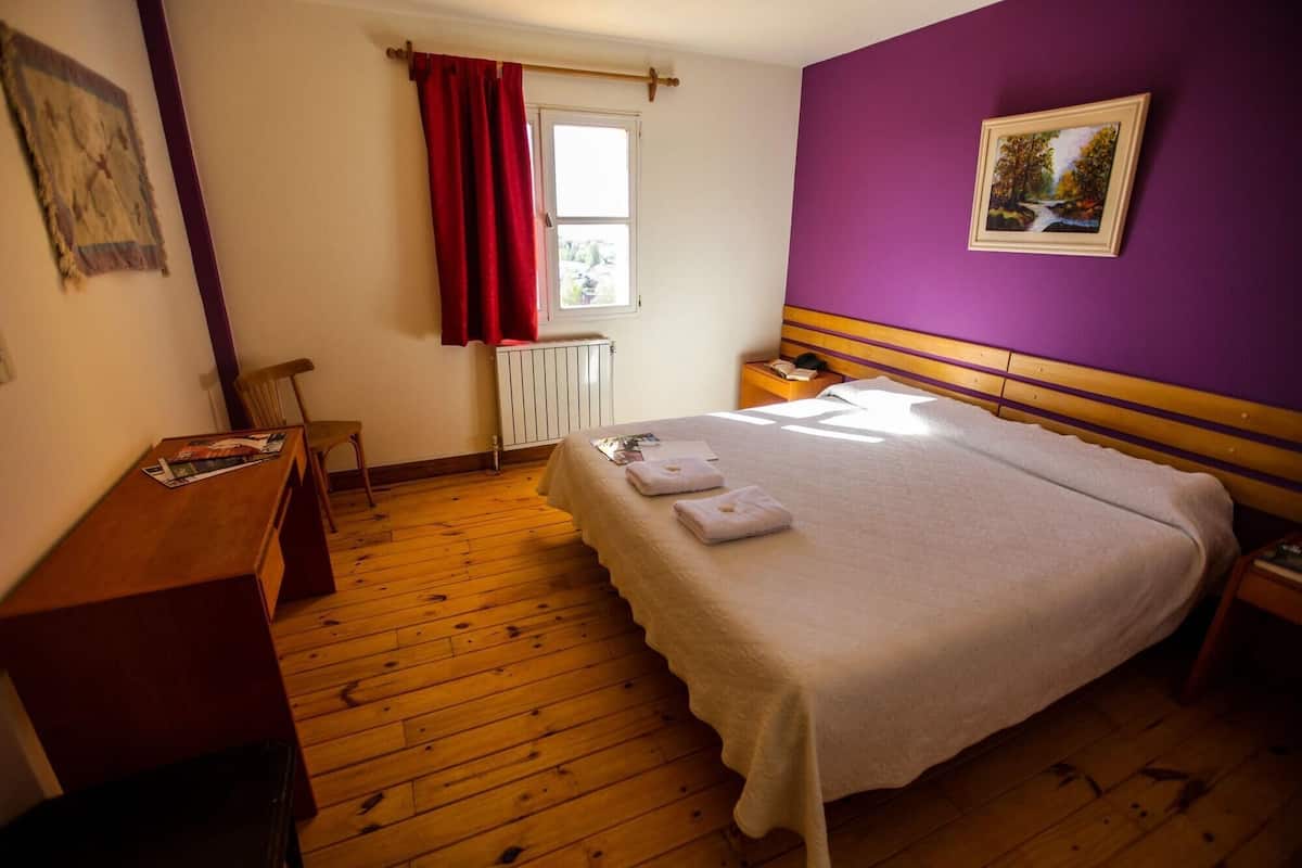 Un dormitorio con cama, suelo de madera, una ventana con cortinas rojas, un radiador, una mesita de noche y un cuadro en la pared.
