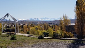 A gazebo with a metal frame and a bench, surrounded by trees and a view of snow-capped mountains.