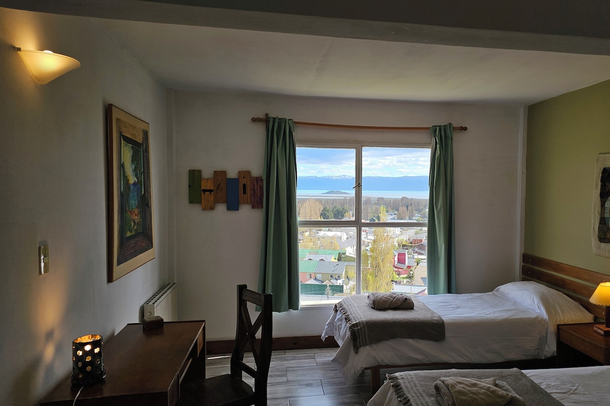 Une chambre d’hôtel avec deux lits, un bureau, une chaise, une fenêtre avec des rideaux et des œuvres d’art accrochées aux murs.
