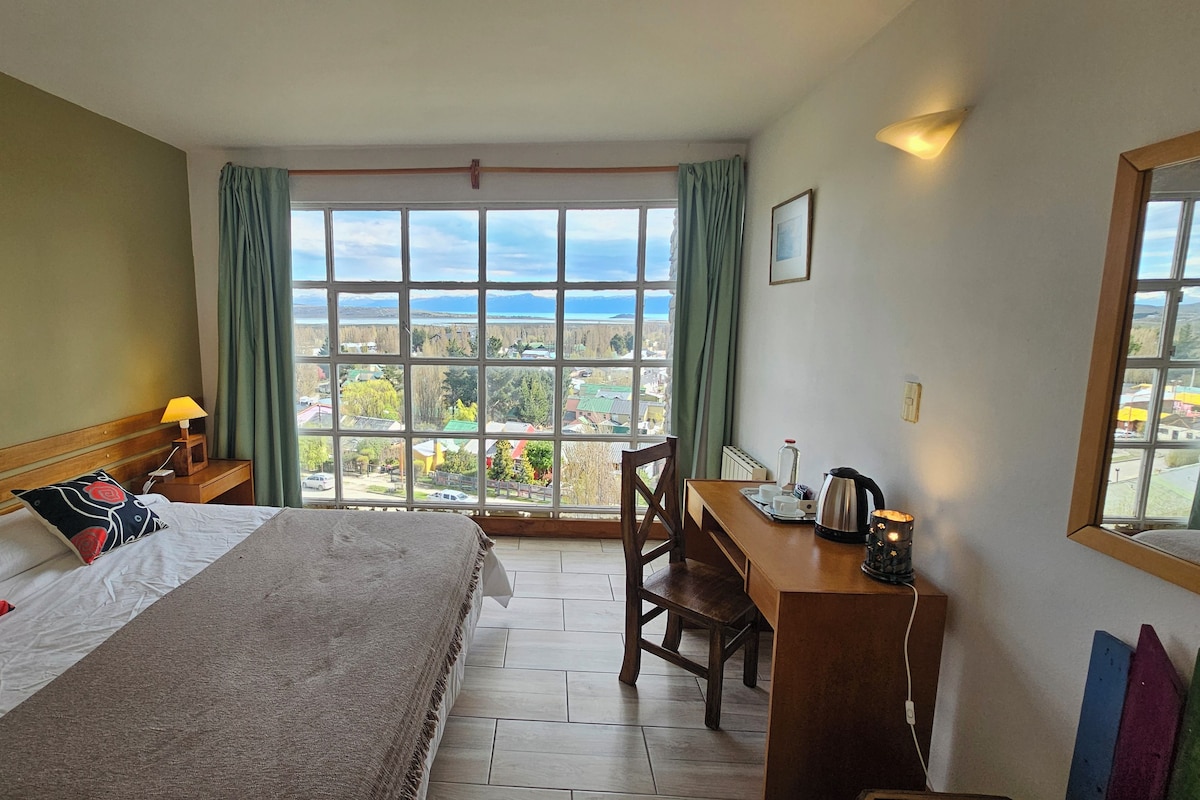 Une chambre d’hôtel avec un lit, un bureau, une chaise, une fenêtre donnant sur un paysage agréable et un tableau au mur.
