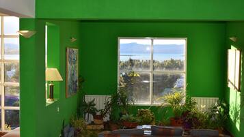 Ein Zimmer mit grünen Wänden, einem großen Fenster mit Blick auf einen See und Berge, einer Couch und verschiedenen topfpflanzengepflanzten Pflanzen.