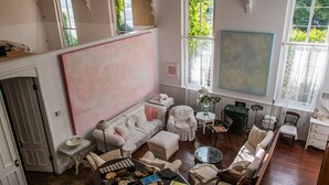 A spacious living room with a large abstract painting, a fireplace, and a view of greenery through the windows.