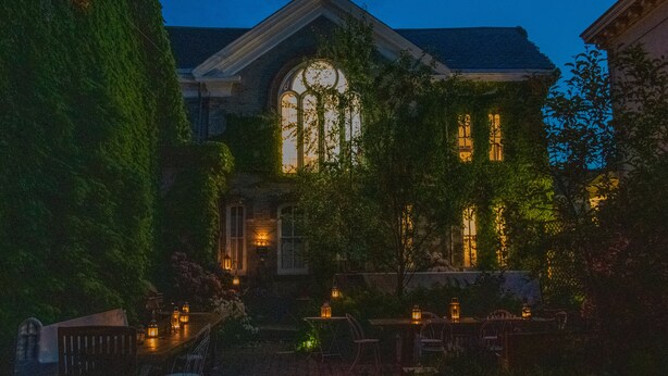 A historic building with a large arched window and ivy-covered walls, surrounded by outdoor seating and lanterns.
