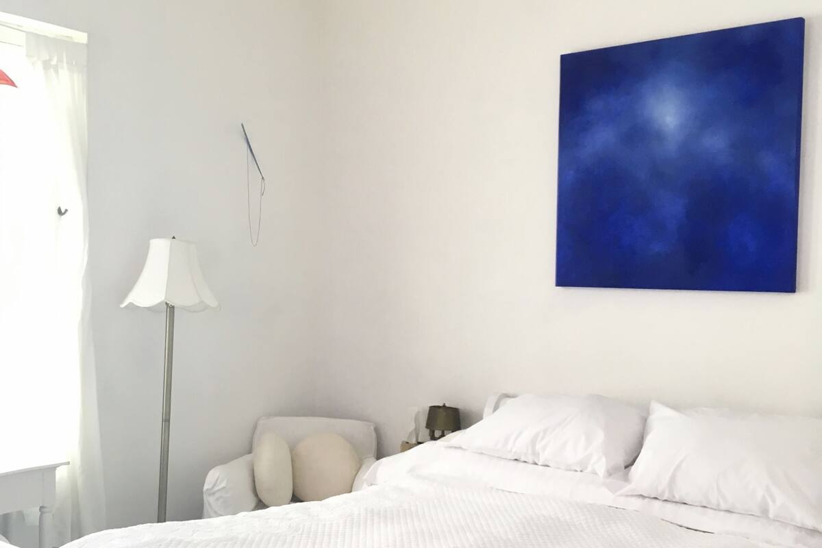 Une chambre à coucher avec un lit, du linge de lit blanc, une lampe sur pied, une table de chevet et un tableau abstrait bleu accroché au mur.