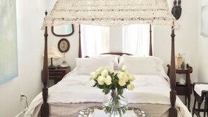 A bedroom with a four-poster bed, a ceiling fan, a vase of white flowers, and a bedside table with a lamp.