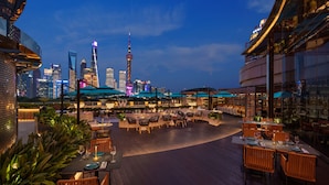 An outdoor dining area with tables and chairs, surrounded by plants and overlooking a city skyline with illuminated buildings.