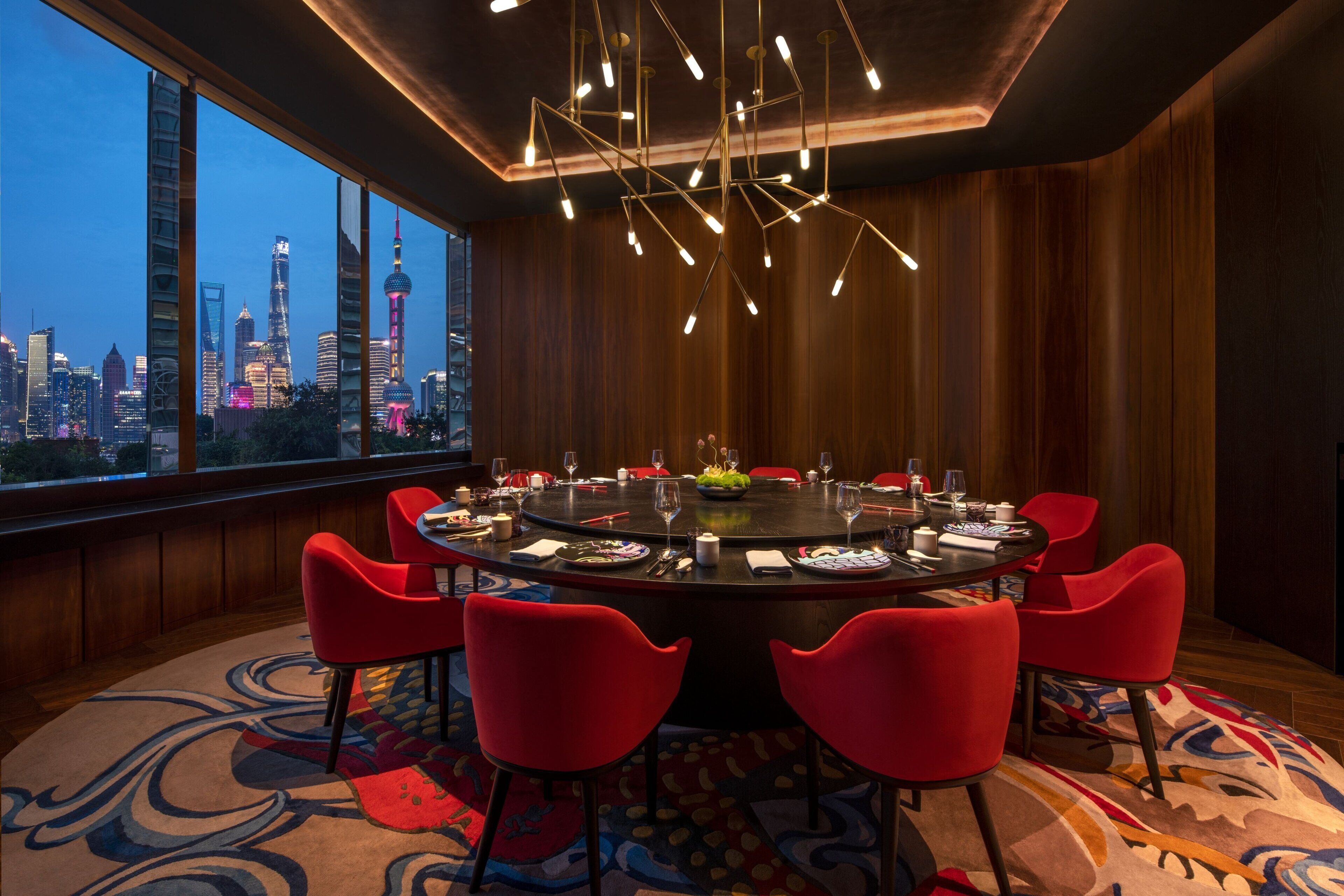 A modern dining room with a round table, red chairs, and a city view.