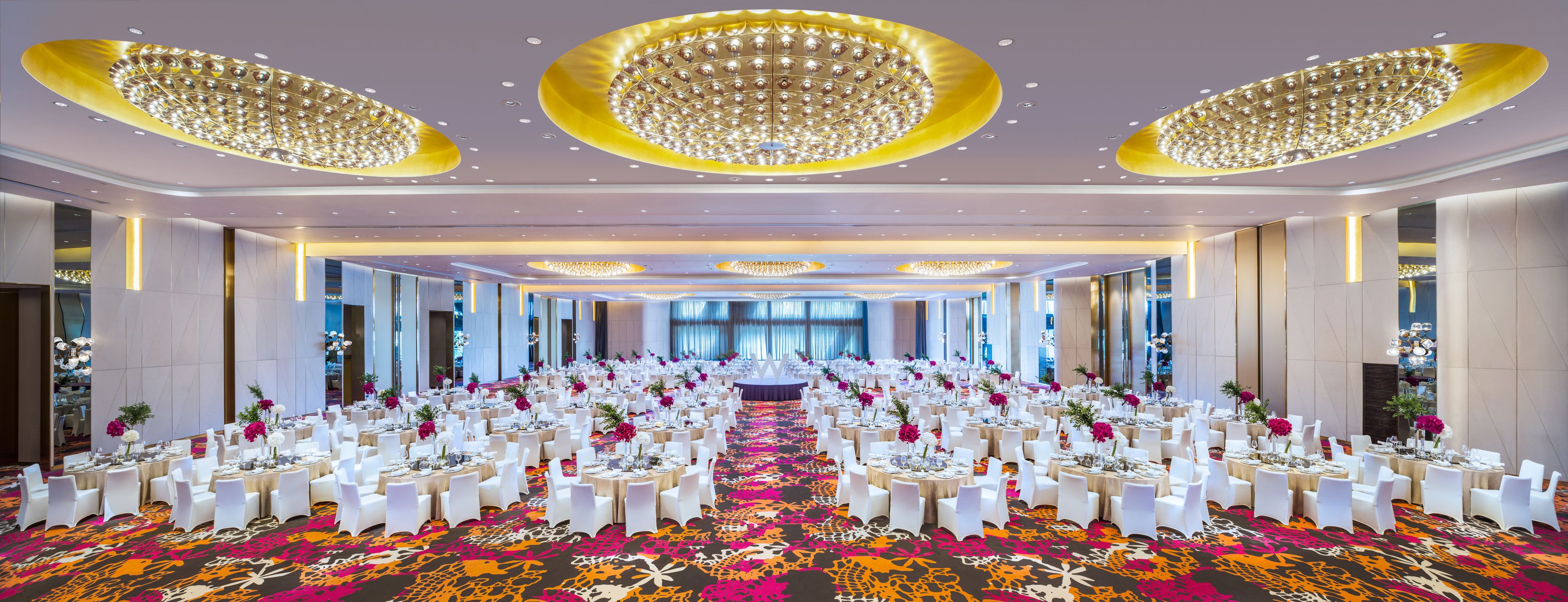 A banquet hall with round tables, white chairs, and floral centerpieces.