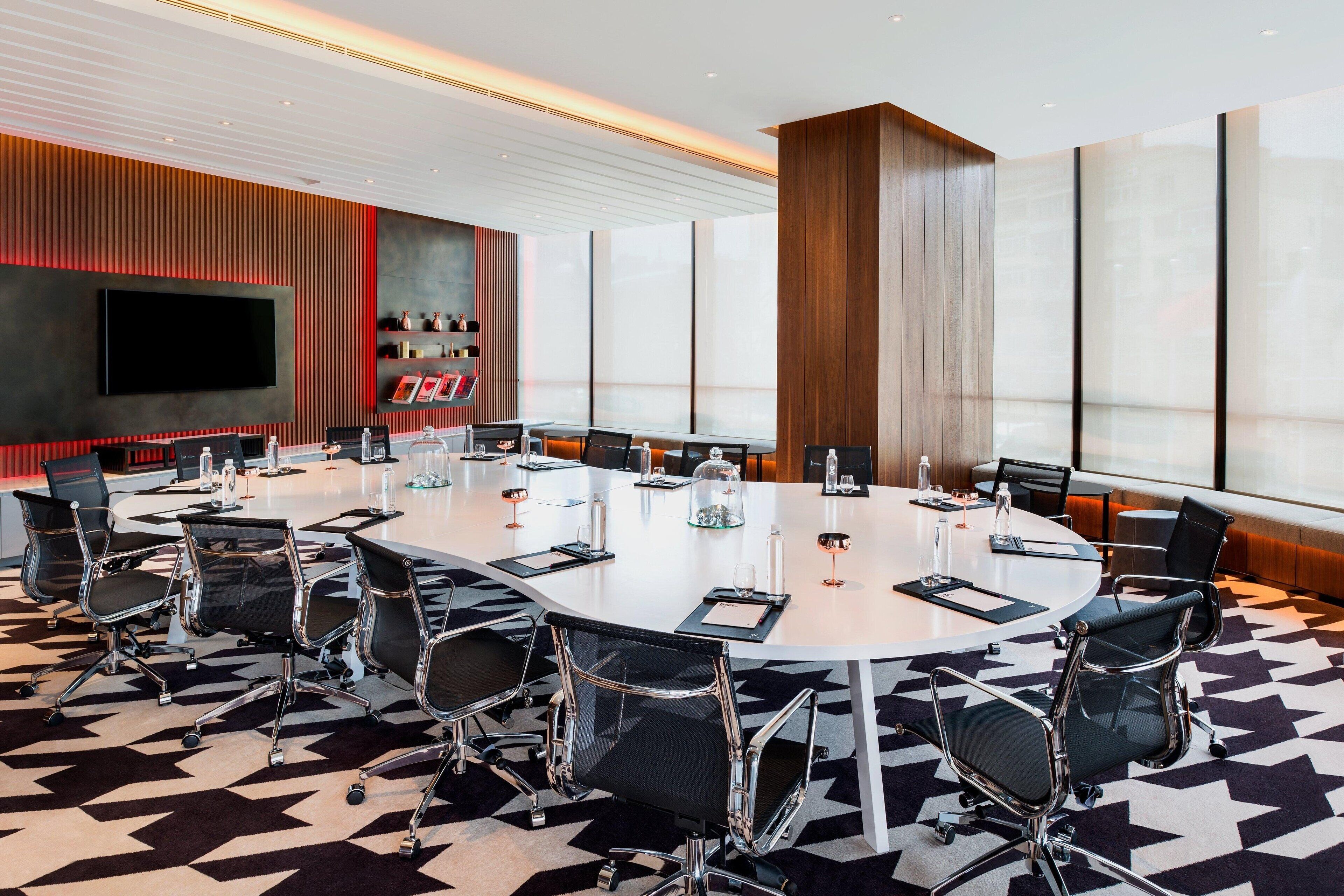A conference room with a large oval table, black chairs, a flat-screen TV, and a red accent wall.