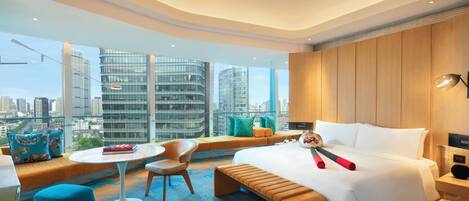 A modern hotel room with a large bed, a seating area with a view of the city, a small table, and a chair.