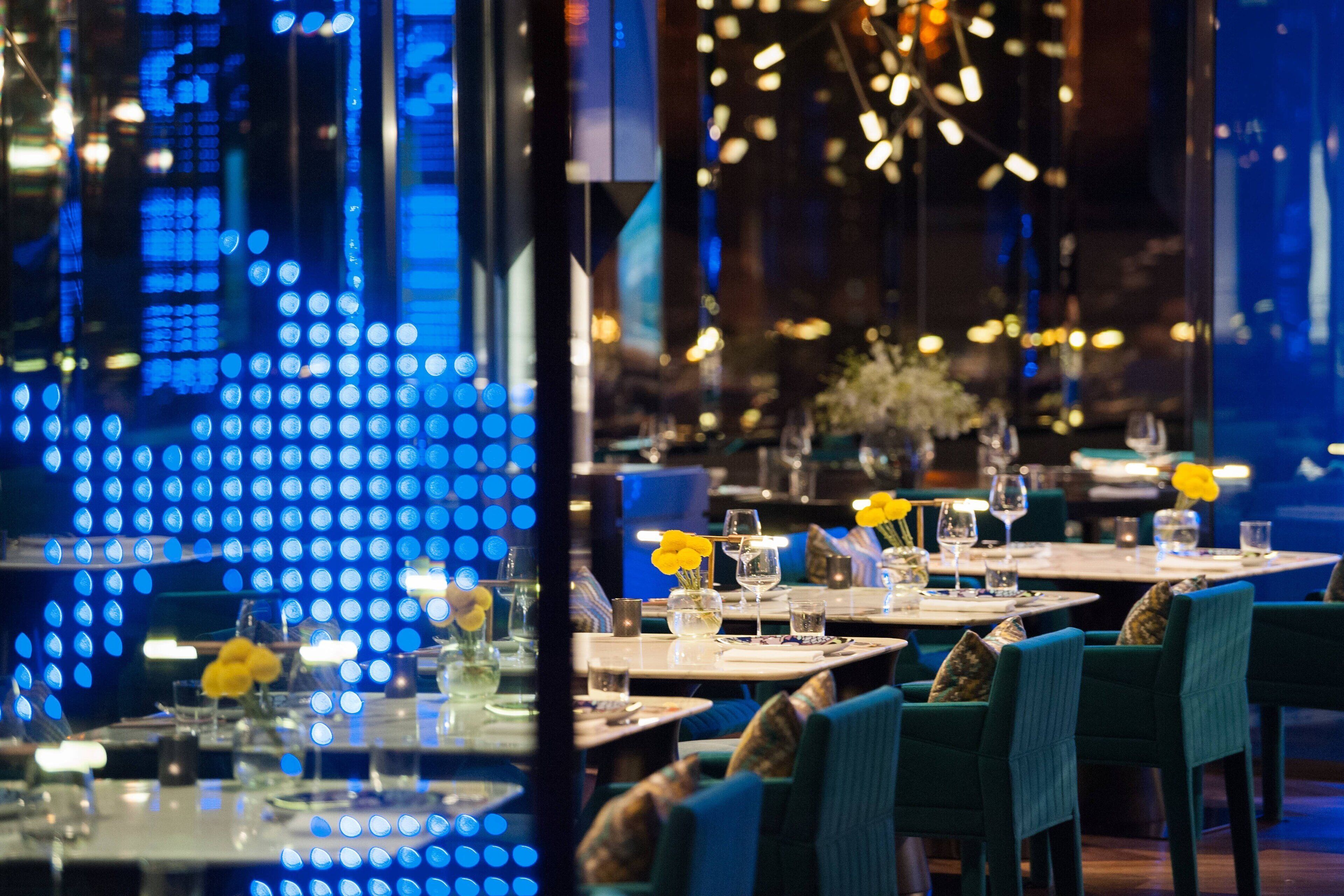 A restaurant with tables set for dining, featuring blue ambient lighting and yellow flowers on the tables.