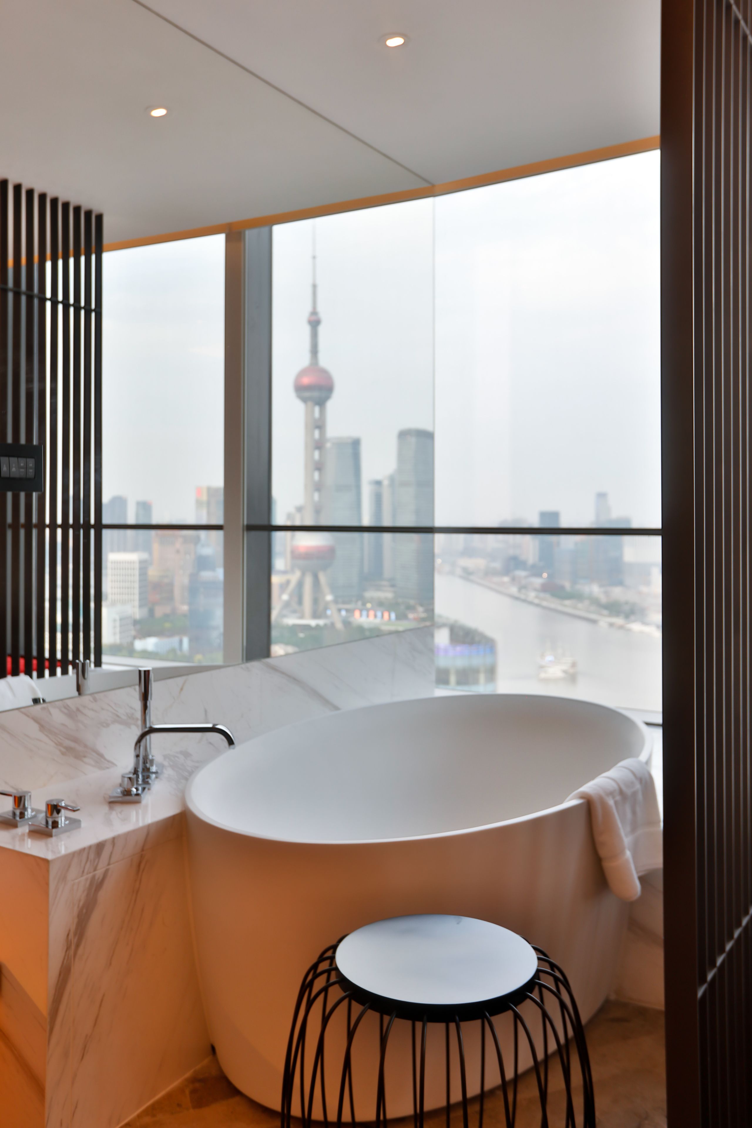 A modern bathroom with a large bathtub, a round stool, and a view of a cityscape through large windows.