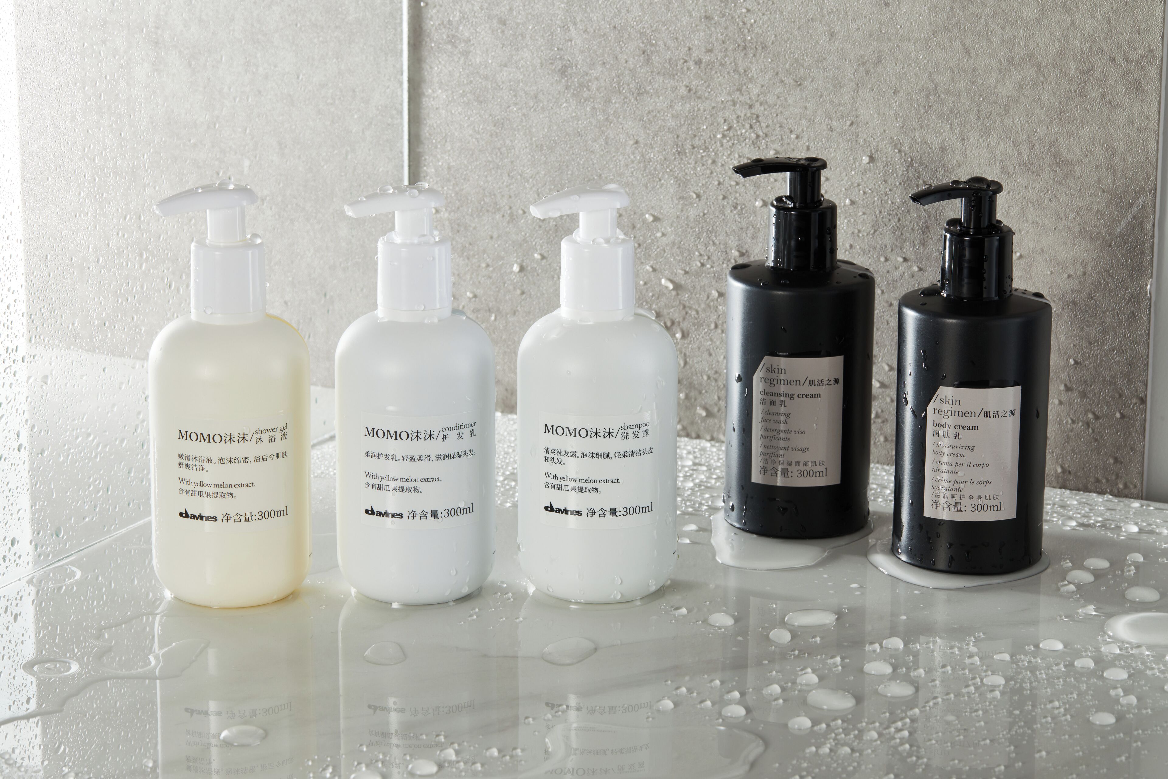 Four bottles of MOMO bath products on a reflective surface with water droplets, against a textured wall.