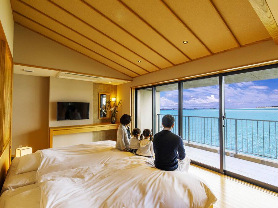 A hotel room with a large bed, a TV, a desk, and a balcony with a view of the ocean.
