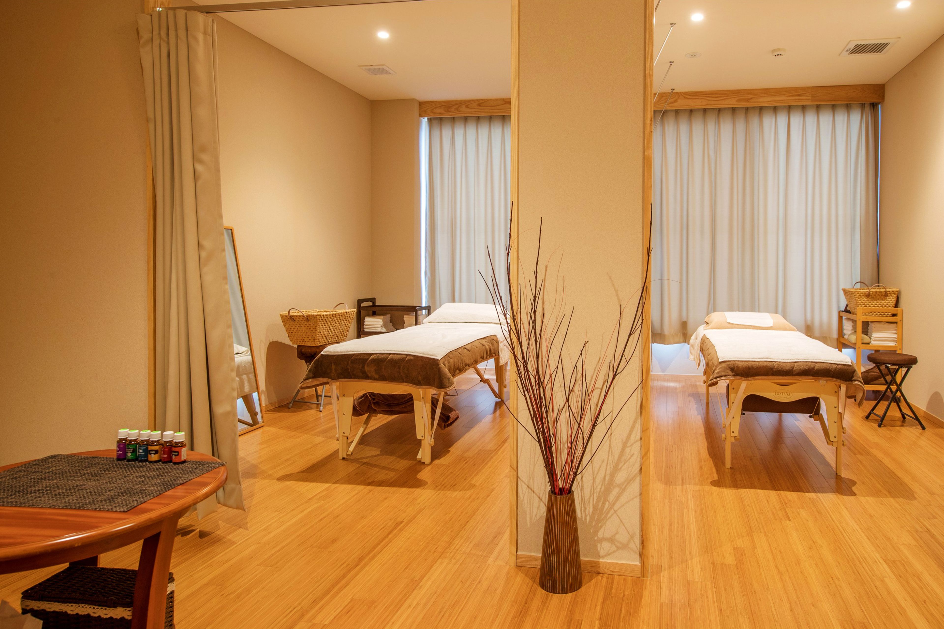 A hotel room with two beds, a wooden floor, a small table with bottles, and a vase with branches.
