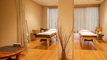 A hotel room with two beds, a wooden floor, a small table with bottles, and a vase with branches.