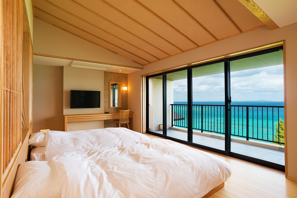 Un dormitorio con una cama grande, un escritorio, un televisor y un balcón con vista al mar.