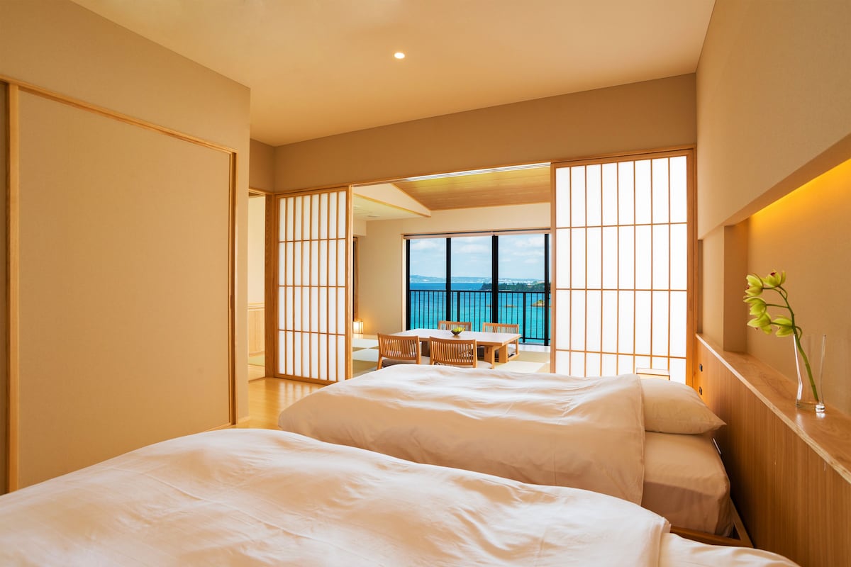 Habitación de hotel con dos camas, zona de comedor con mesa y sillas, y vista al mar a través de amplios ventanales.