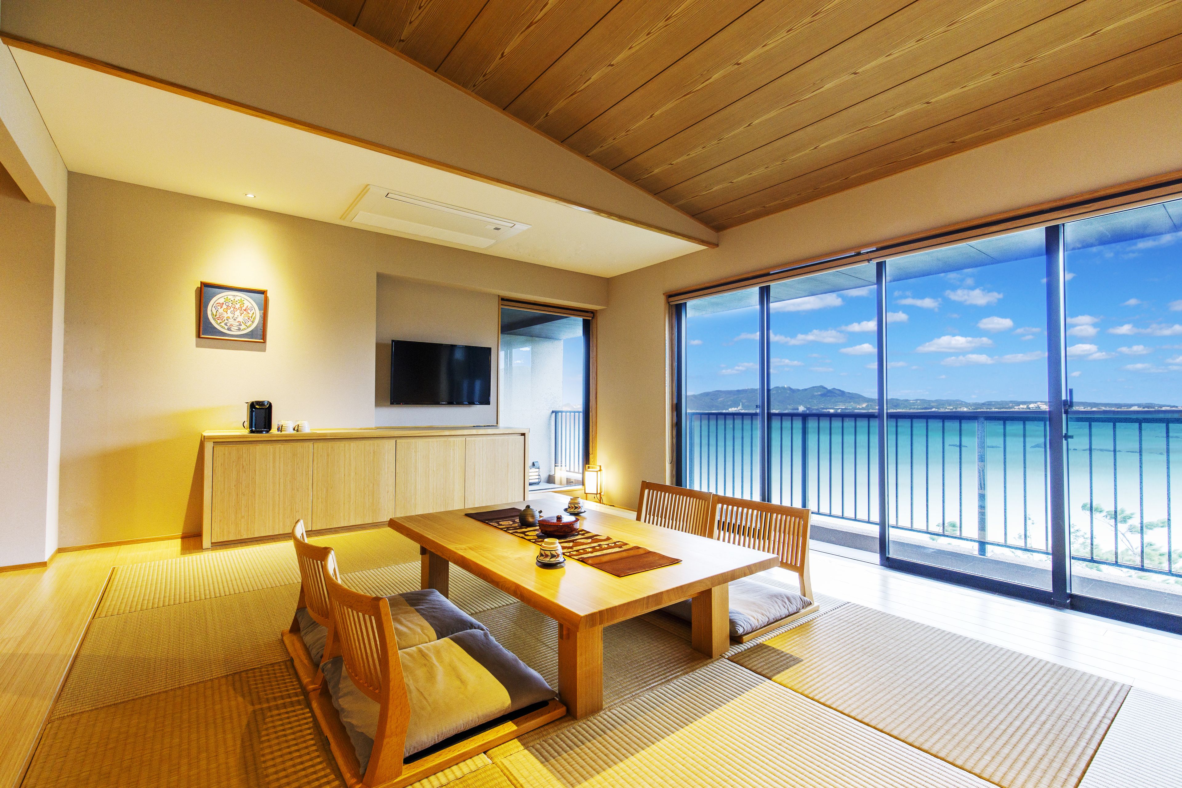 A modern dining area with a wooden table and chairs, a television, and a view of the sea.