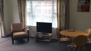 Una habitación con una televisión de pantalla plana sobre un pedestal, un sillón marrón, una mesa redonda con cuatro sillas y una ventana con cortinas.