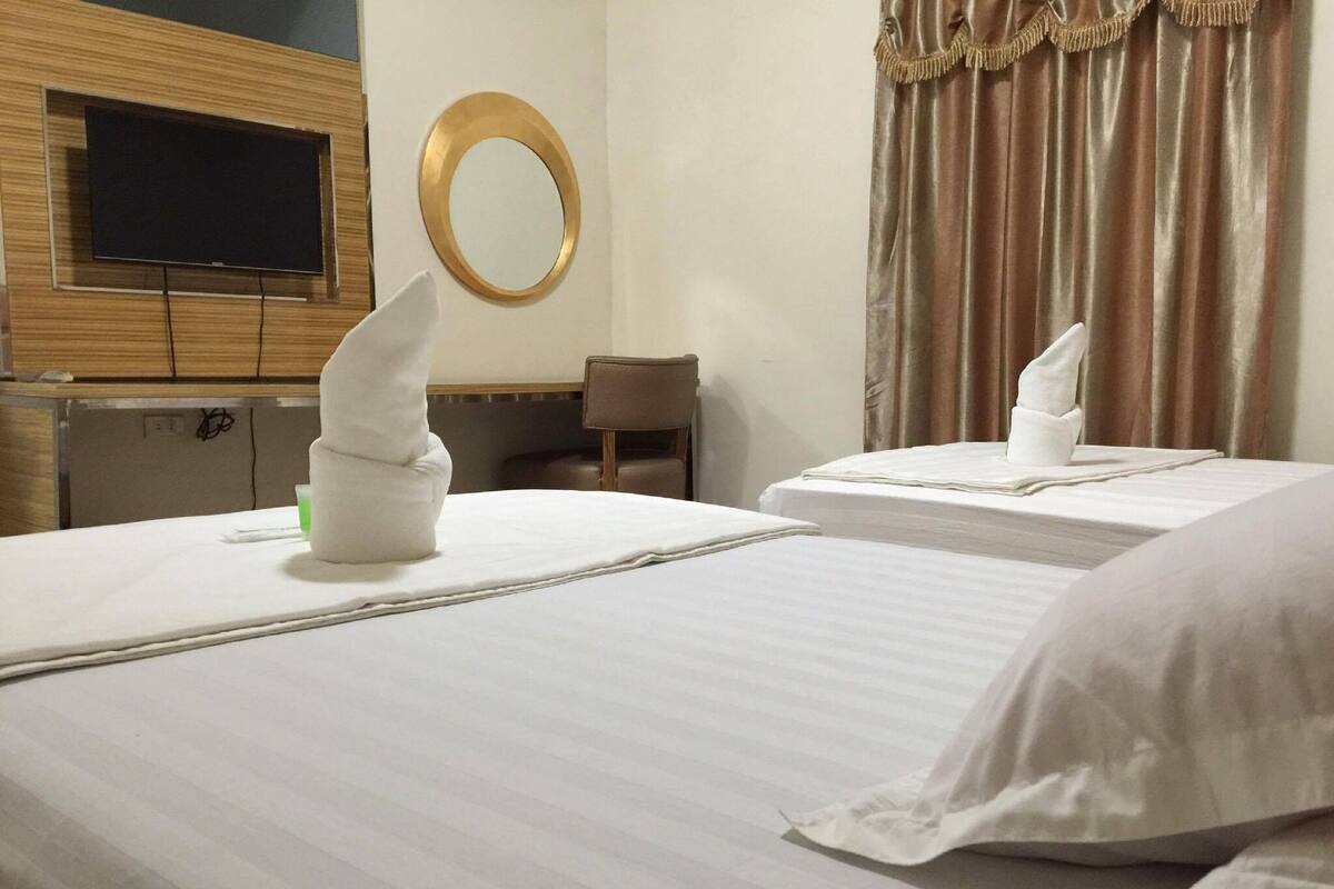 A hotel room with two beds, a flat-screen TV mounted on a wooden panel, a round mirror, and a window with curtains.