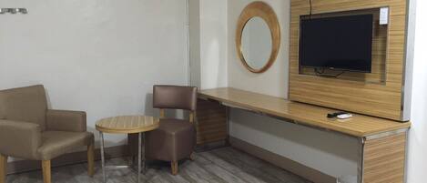 A hotel room with a sofa, a small round table, a chair, a TV mounted on a wooden cabinet, and an air conditioning unit on the wall.