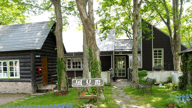 Una cabaña de troncos con un porche de madera, un letrero en idioma extranjero y un jardín con cerca blanca a cuadros.
