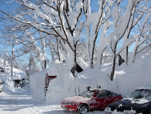 A red car parked in a snowy area with trees and a building in the background. - Woody House Kazamidori (Myoko)