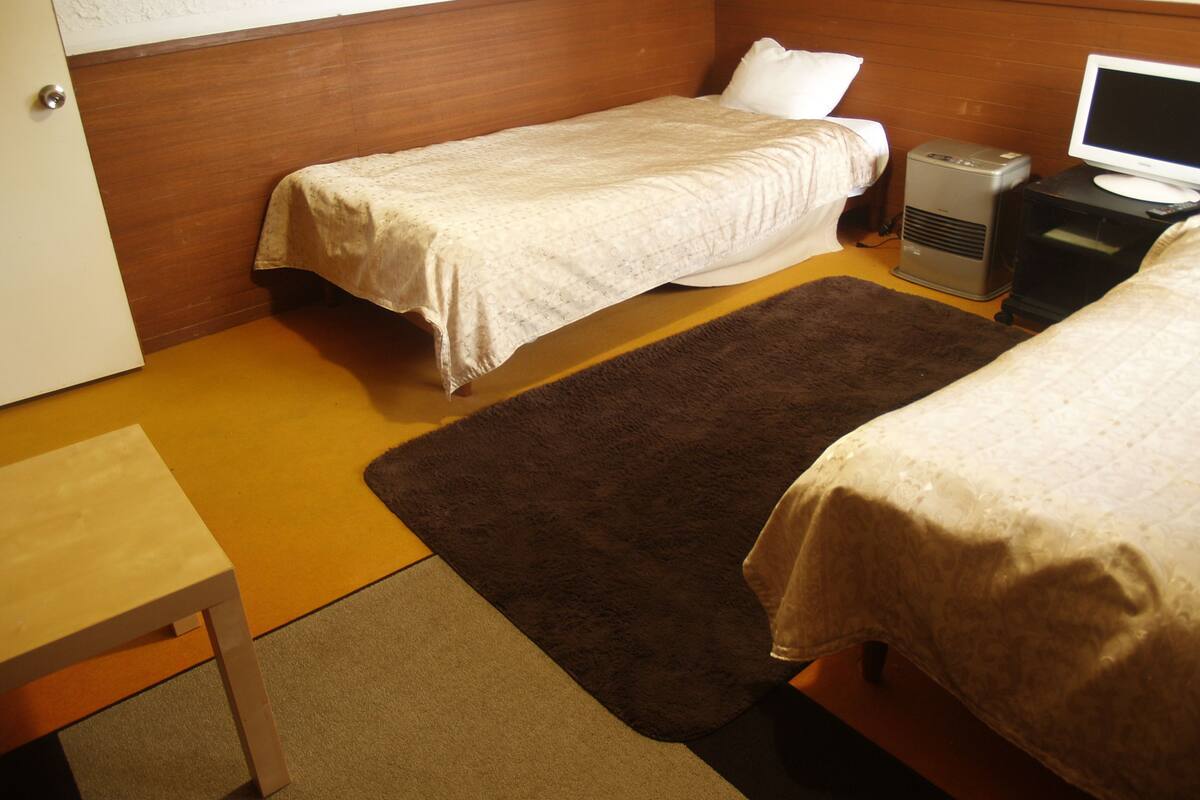 A small, compact room with two single beds, a desk, a computer, and a heater.