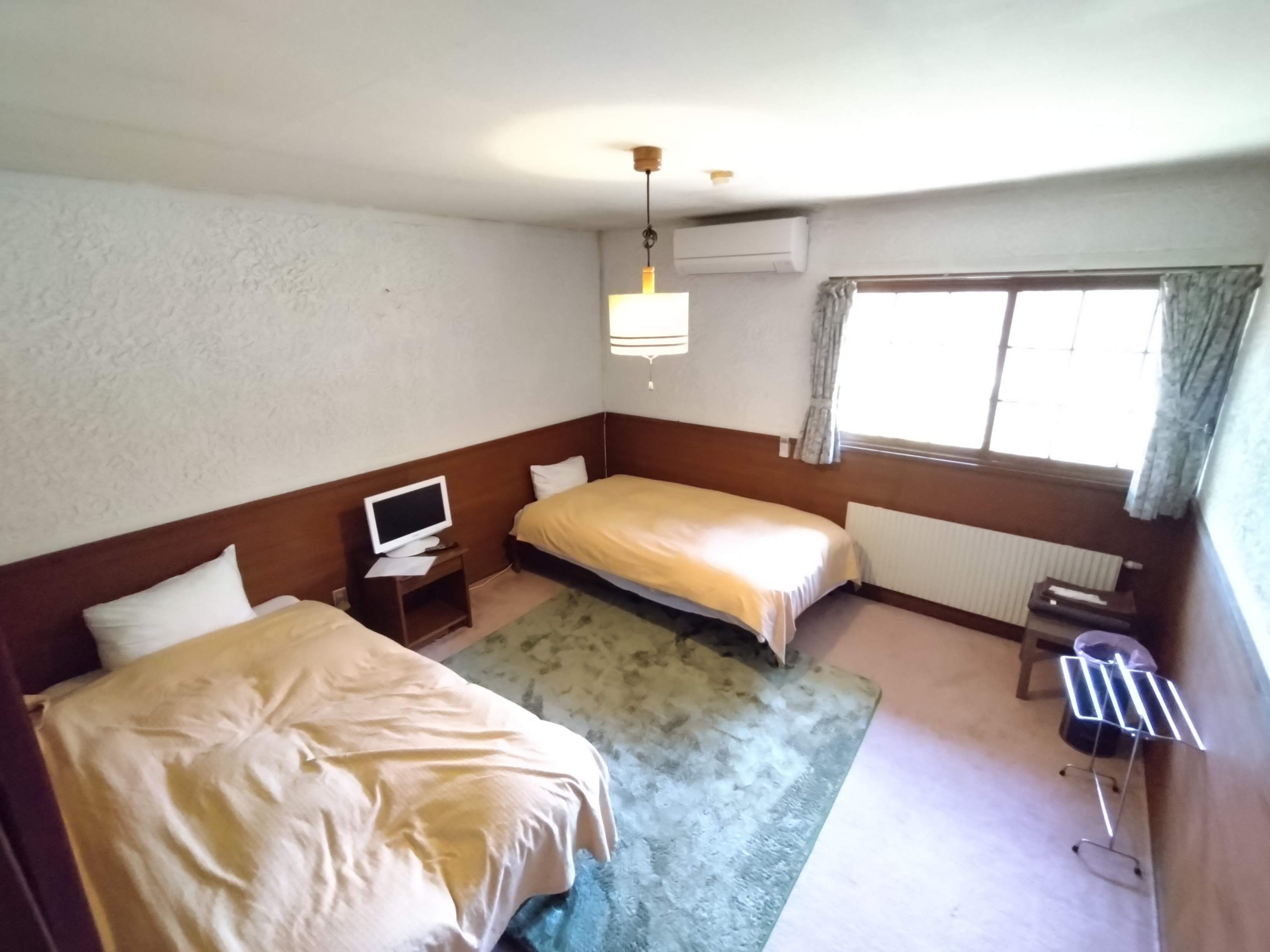 A hotel room with two beds, a TV on a side table, a radiator, and a window with curtains.