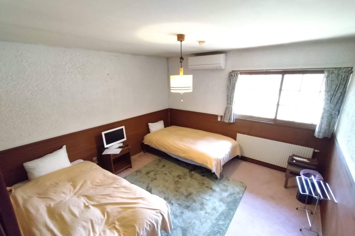 A hotel room with two beds, a TV on a side table, a radiator, and a window with curtains.