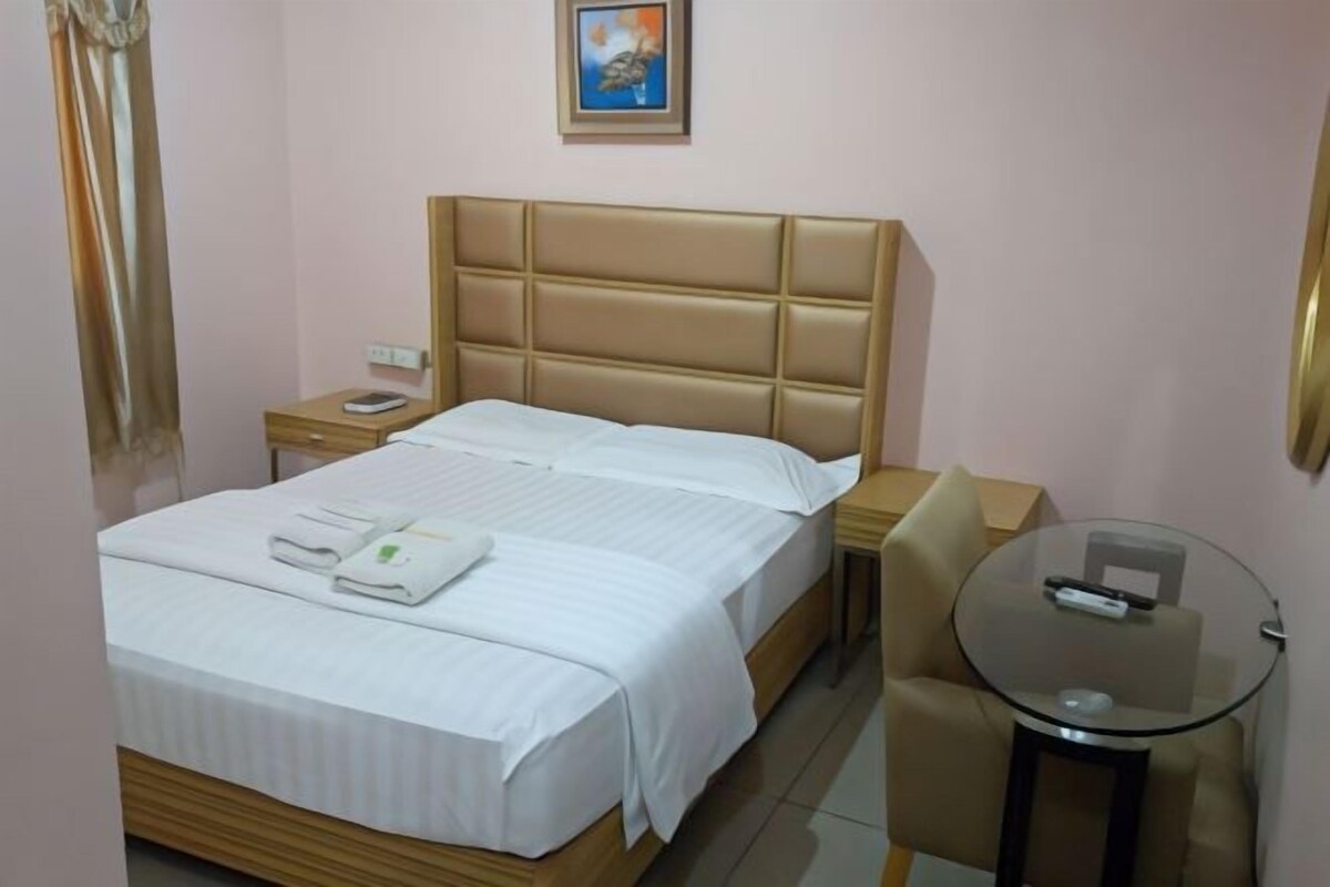 A neatly made bed with white linens and a folded towel, a bedside table with a phone, a small round table with a remote control, and a framed picture on the wall.