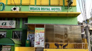 Exterior del Hotel Meaco Royal con fachada verde y amarilla, una escalera visible que conduce a la entrada y un cartel que anuncia las tarifas de las habitaciones.