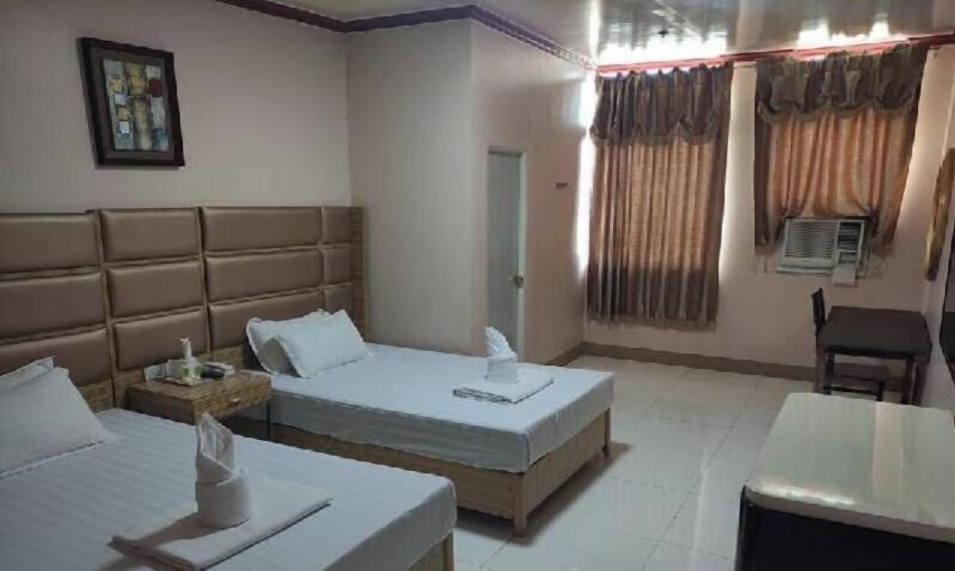 Habitación de hotel con dos camas, una mesita de noche con lámpara, una silla, una pequeña mesa y una ventana con cortinas.
