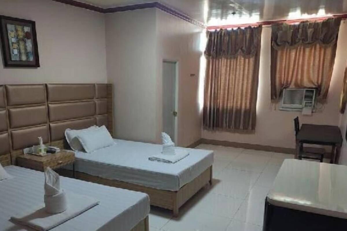 Habitación de hotel con dos camas, una mesita de noche con lámpara, una silla, una pequeña mesa y una ventana con cortinas.
