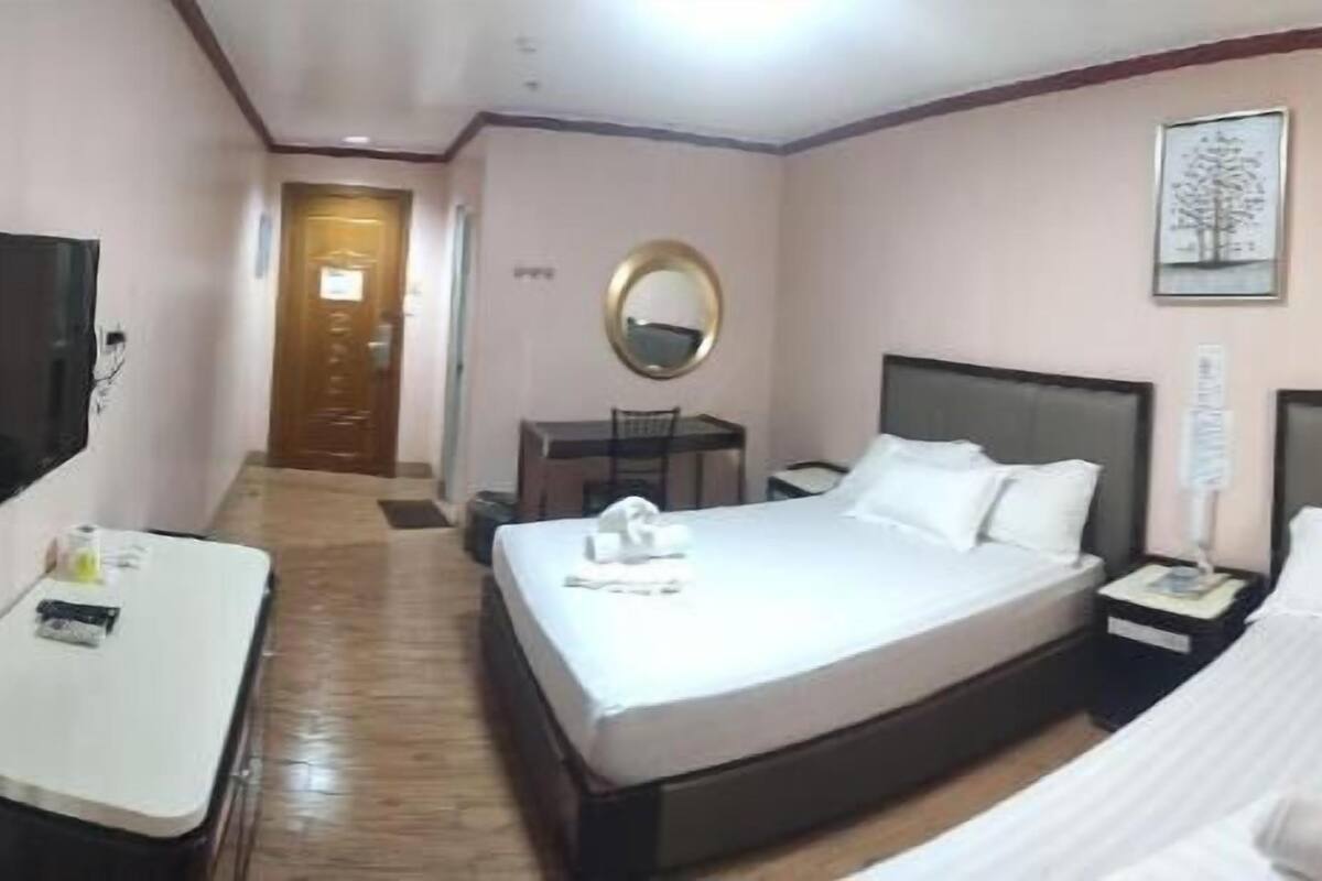 Una habitación de hotel con cama, mesita de noche, un escritorio con silla, televisión y espejo.